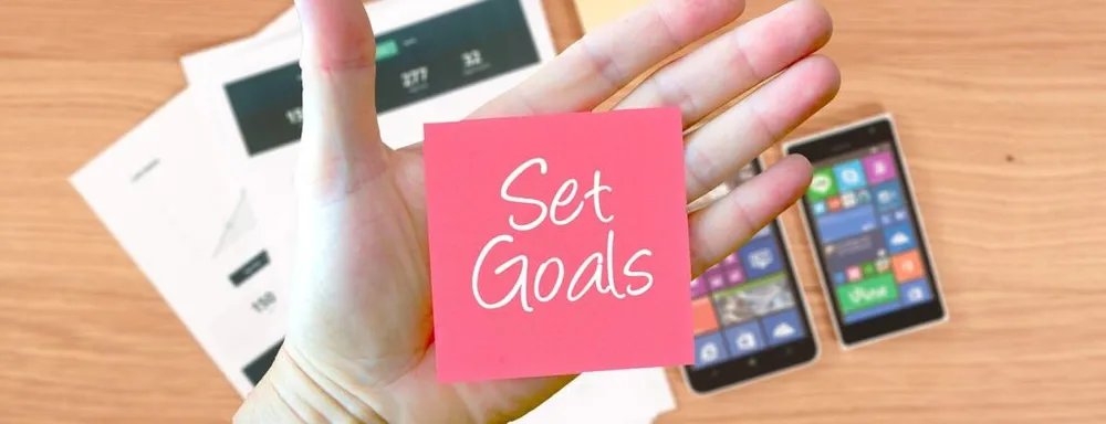 A hand holding a pink sticky note that says Set Goals